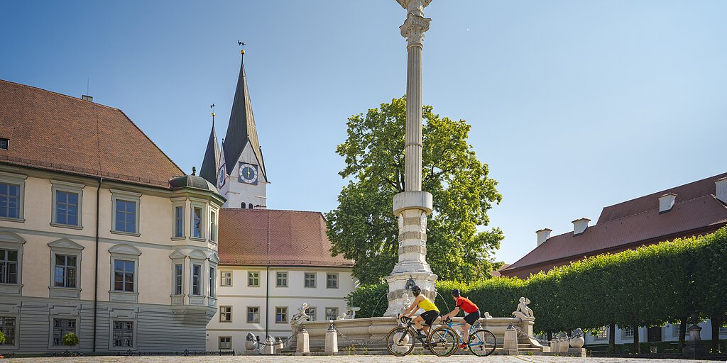 Zwei Radfahrer fahren an einer Mariensäule auf einem Platz mit historischen Gebäuden vorbei.
