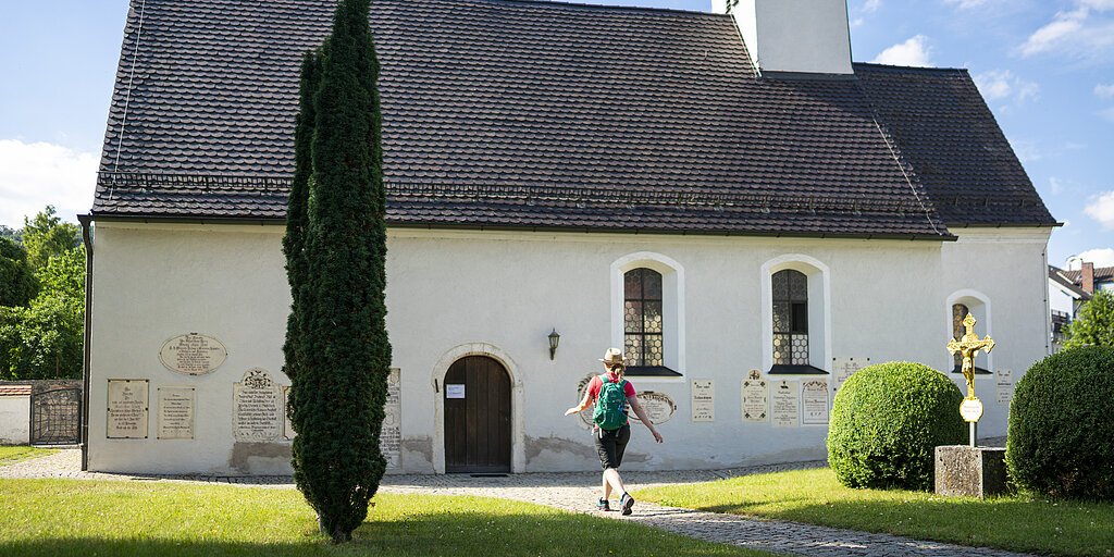 Person mit Rucksack geht auf eine kleine Kirche mit Zwiebelturm zu, umgeben von Bäumen und Sträuchern.