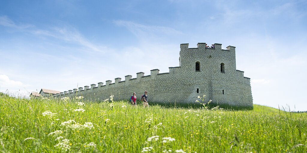 Zwei Wanderer gehen an einer historischen Steinmauer auf einer grünen Wiese unter blauem Himmel vorbei.