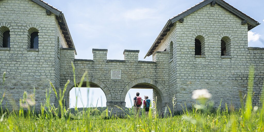 Zwei Personen mit Rucksäcken stehen unter einem steinernen Torbogen einer historischen Anlage bei sonnigem Himmel.