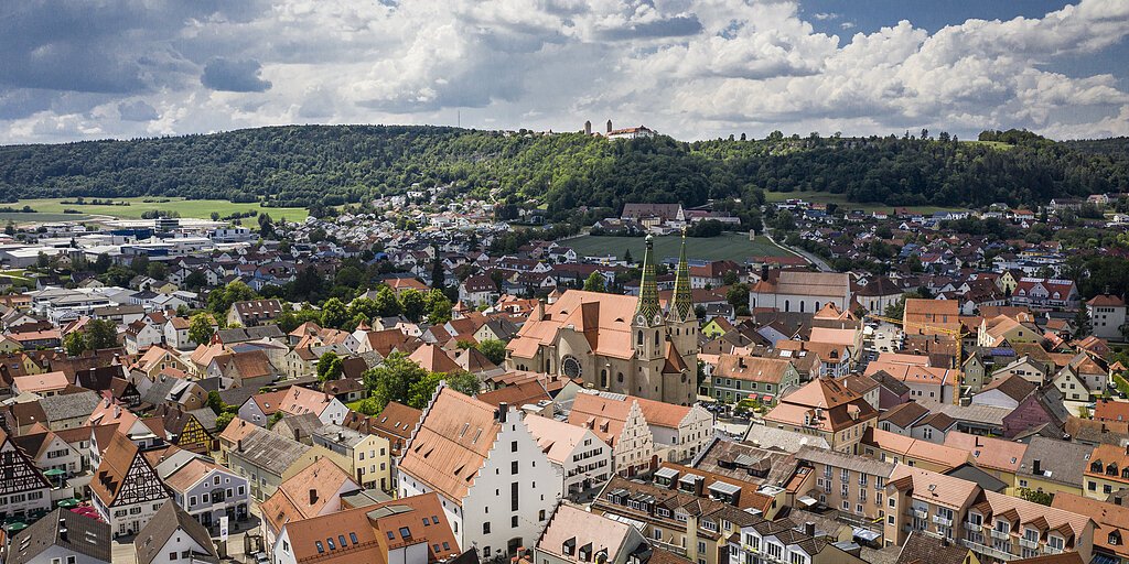 Luftaufnahme einer Stadt mit roten Dächern, Kirche mit zwei Türmen und bewaldetem Hügel im Hintergrund unter bewölktem Himmel.