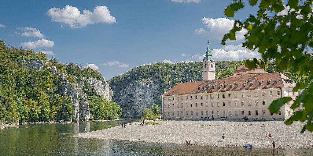 Flussufer mit Kiesstrand, altes Gebäude mit Turm und bewaldete Felsen unter blauem Himmel mit Wolken