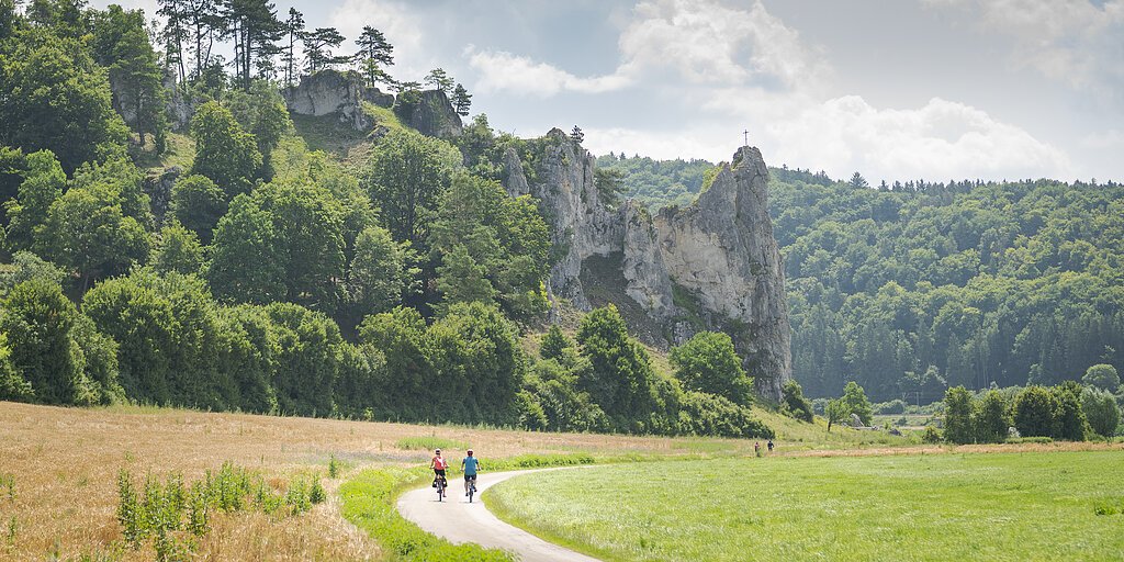 Zwei Radfahrer auf einem Weg vor bewaldeten Felsen mit einem Kreuz auf dem höchsten Felsen.