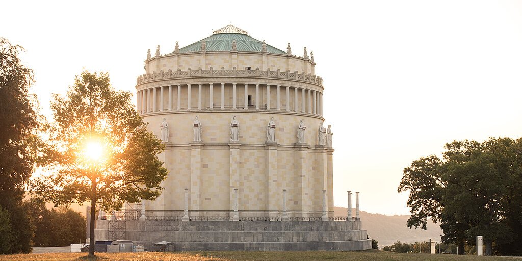 Rundes historisches Gebäude mit Statuen auf dem Sims, Sonne hinter Baum im Park bei Abendlicht