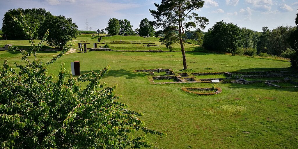 Grüne Wiesen mit Baum, Ruinenfundamenten und blauem Himmel mit Wolken im Hintergrund.