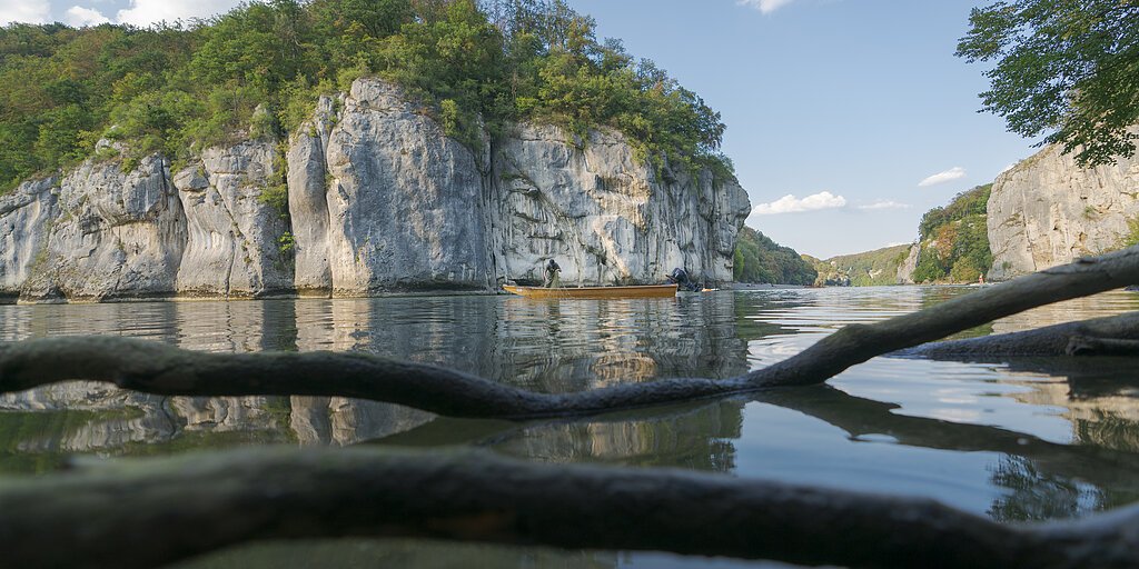 Fluss mit Felsen und Bäumen, Boot mit zwei Personen, Äste im Vordergrund im Wasser sichtbar