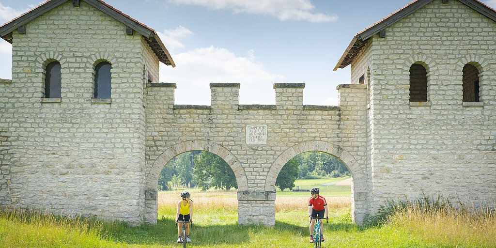Zwei Radfahrer fahren durch zwei Bögen einer historischen Steinmauer in einer grünen Wiesenlandschaft.
