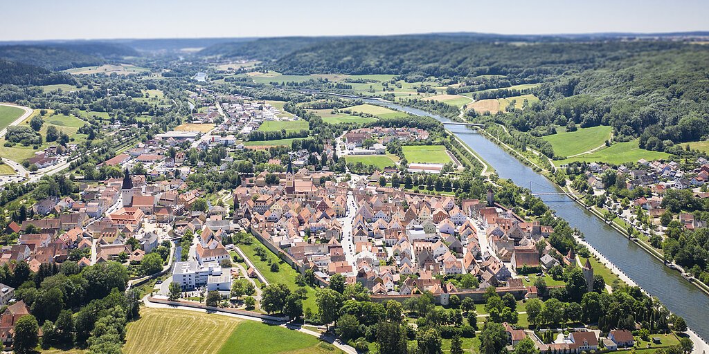 Luftaufnahme einer Kleinstadt mit Fluss, umgeben von Feldern und Wäldern unter blauem Himmel.