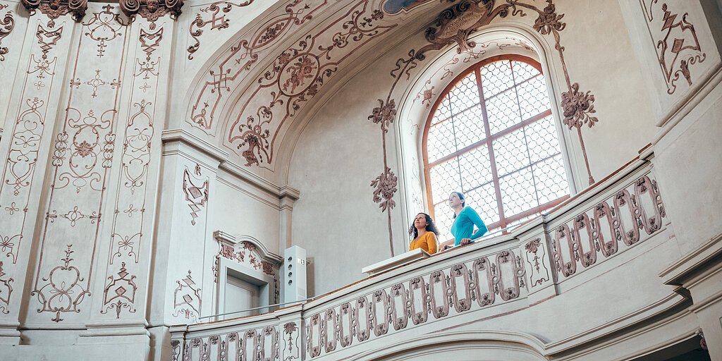 Zwei Personen stehen auf einem verzierten Balkon in einem historischen, mit Fresken geschmückten Innenraum.