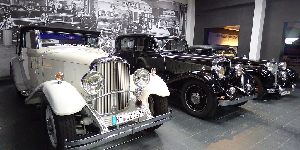 Drei Oldtimer-Autos nebeneinander in einem Museum mit historischem Schwarz-Weiß-Foto im Hintergrund.
