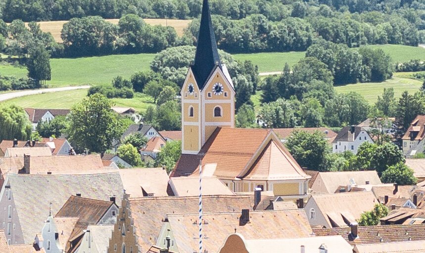 Blick auf eine Stadt mit roten Dächern, Flussufer, Bäumen und bewaldeten Hügeln im Hintergrund bei klarem Himmel.